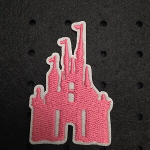 Könnte beinhalten: Gestickter rosa Burg-Aufnäher mit weißem Rand. Der Aufnäher zeigt ein detailliertes Design eines Märchenschlosses mit mehreren Türmen und Fahnen. Der Aufnäher ist wahrscheinlich für Kleidung oder Accessoires.