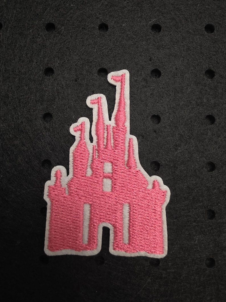 Op de afbeelding: Roze geborduurde kasteelpatch met een witte rand. De patch heeft een gedetailleerd ontwerp van een sprookjeskasteel met meerdere torentjes en vlaggen. De patch is waarschijnlijk voor kleding of accessoires.