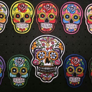 Könnte beinhalten: Eine Sammlung farbenfroher Totenkopf-Patches mit floralen Designs. Die Patches weisen verschiedene Farben auf, darunter Gelb, Rot, Rosa, Blau, Grün und Lila, mit aufwendigen Details. Die Totenköpfe haben eine Sugar-Skull-Ästhetik, perfekt für Feierlichkeiten zum Tag der Toten.