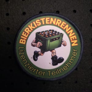 Könnte beinhalten: Runder Aufnäher mit dem Text "BIERKISTENRENNEN Lizenzierter Teilnehmer" in einem grünen und orangen Design. Der Aufnäher zeigt eine Cartoonfigur, die eine grüne Kiste voller Bierflaschen trägt.
