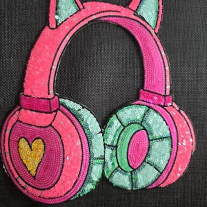 Könnte beinhalten: Ein pinkfarbener Kopfhörer-Applikation mit Katzenohren. Die Ohrmuscheln sind pink und türkis, mit einem goldenen Herzdetail auf einer Ohrmuschel. Das Stirnband und die Ohrmuscheln sind schwarz umrandet.