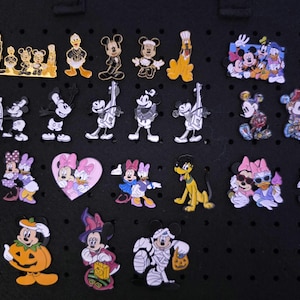 Könnte beinhalten: Eine Sammlung von achtundzwanzig Emaille-Anstecknadeln mit verschiedenen Disney-Figuren, darunter Mickey Maus, Minnie Maus, Donald Duck, Daisy Duck und Pluto in unterschiedlichen Posen, Outfits und Farben, angeordnet auf einer schwarzen Pinnwand.