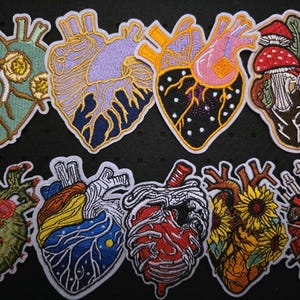 Könnte beinhalten: Eine Sammlung von aufgestickten Aufnähern mit anatomischen Herzmotiven. Jeder Aufnäher zeigt ein einzigartiges Farbschema und Design, darunter Blumen-, Pilz- und Himmelsmotive. Die Aufnäher sind mit einem weißen Rand umrandet.