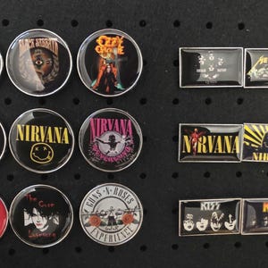 Könnte beinhalten: Eine Sammlung runder und rechteckiger Band-Anstecknadeln mit ikonischen Rockbands wie Nirvana, Kiss und Black Sabbath. Die Anstecknadeln haben glänzende Oberflächen und leuchtende Farben, mit Bandlogos und Albumcovern. Die Anstecknadeln sind auf einer schwarzen Lochplatte angeordnet.