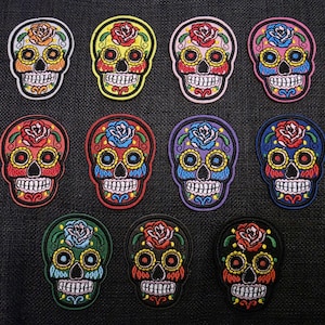 Könnte beinhalten: Eine Sammlung von elf bunten, gestickten Totenkopf-Patches. Jeder Patch zeigt ein detailliertes Sugar-Skull-Design mit einer Rose oben, in verschiedenen Farben wie Rot, Gelb, Blau, Rosa und Grün. Die Patches befinden sich auf einem dunklen Hintergrund.