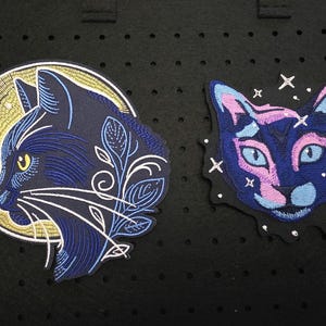 Könnte beinhalten: Zwei gestickte Katzenaufnäher. Einer zeigt ein marineblaues Katzenprofil mit gelbem Mondhintergrund und weißen floralen Akzenten. Der andere Aufnäher hat ein blaues und rosa Katzengeischt mit Sternakzenten. Beide Aufnäher befinden sich auf schwarzem Hintergrund.