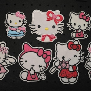 Könnte beinhalten: Eine Sammlung von acht gestickten Aufnähern mit einer Cartoon-Figur. Jeder Aufnäher zeigt die Figur in verschiedenen Posen, geschmückt mit Schleifen, Kleidern und Accessoires in Rosa, Blau und Weiß, vor einem schwarzen Hintergrund.