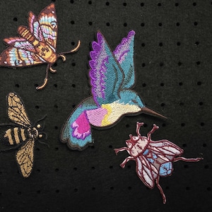 Könnte beinhalten: Gestickte Aufnäher mit einem Schmetterling, einem Kolibri, einer Biene und einem Käfer. Die Aufnäher zeigen leuchtende Farben und detaillierte Stickereien. Der Schmetterling hat braune, blaue und orangefarbene Flügel. Der Kolibri hat blaue und violette Flügel.