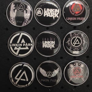 Könnte beinhalten: Neun runde Linkin Park Band-Anstecknadeln mit verschiedenen Logos und Texten. Die Anstecknadeln sind hauptsächlich schwarz und weiß, einige mit roten Akzenten. Designs beinhalten das Logo der Band, Flügel und den Bandnamen.