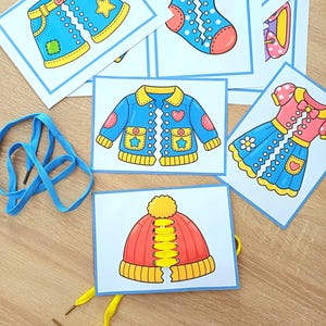 Könnte beinhalten: Ein Set aus Lernkarten mit farbenfrohen Illustrationen von Kleidungsstücken wie Shorts, einer Socke, einer Jacke, einem Kleid und einer Mütze, ergänzt durch blaue und gelbe Schnürsenkel für Schnürübungen durch die Ausstanzungen der Karten.