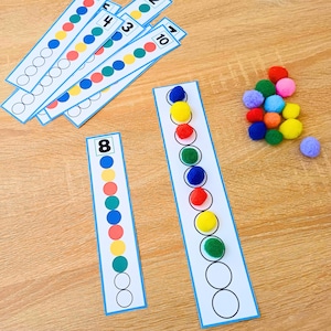 Puede incluir: Un conjunto de tiras educativas para contar con los números del 1 al 10 y círculos de colores correspondientes. Algunos círculos están rellenos con pompones difusos a juego en rojo, azul, verde, amarillo, naranja, morado y rosa, otros vacíos.