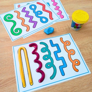 May include: Two white activity sheets featuring colorful printed lines in various shapes including zigzags, swirls, and curves. One sheet has yellow, magenta, teal, blue, and orange modeling clay shaped to match the printed lines. A blue clay ball and a yellow lidded container are present.