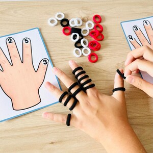 May include: A learning activity set featuring two printed cards showing illustrations of hands with numbered fingers, accompanied by a collection of small, circular elastic hair ties in black, white, and red colors for counting practice.