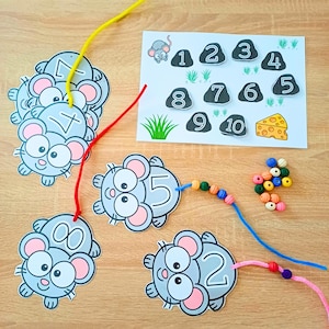 May include: A children's educational counting activity set featuring grey mouse-shaped cards with numbers, a white reference sheet with numbered stones and a cheese illustration, colourful plastic lacing strings, and small assorted plastic beads.