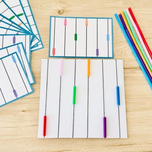 Könnte beinhalten: Ein Set aus weißen rechteckigen Karten mit vertikalen schwarzen Linien und kleinen farbigen Kunststoffzylindern, die an den Linien befestigt sind. Die Farben umfassen Rot, Orange, Gelb, Grün, Blau, Lila und Pink. Eine Sammlung passender farbiger Stifte ist enthalten.