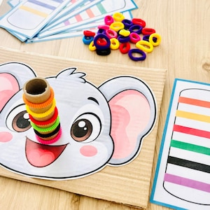 May include: A cardboard activity board featuring a cartoon elephant face with a vertical cardboard tube attached. A stack of small, colorful elastic hair ties in orange, yellow, red, green, black, and pink are placed on the tube. Includes matching pattern cards.