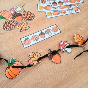 Peut inclure: Une guirlande composée de découpes de citrouilles orange, de glands marron, de pommes de pin, de champignons rouges à pois blancs et de feuilles d'automne jaune-orange sur un ruban noir, avec des découpes en papier et des bandes assorties.