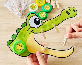 May include: A green and yellow crocodile-shaped cutout with a large eye and a number 4 bottle cap attached. The set includes several wooden clothespins and additional colorful bottle caps labeled with numbers 7, 9, 13, and 20 in a white bag.