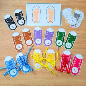 Puede incluir: Un conjunto de tarjetas educativas con forma de zapato en varios colores, incluyendo negro, naranja, morado, verde, azul, amarillo, rosa y gris. Cada tarjeta está etiquetada como zapato izquierdo o derecho. Una tarjeta guía muestra dos huellas.