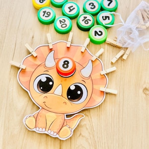 Pode incluir: Um cartão de aprendizagem em forma de dinossauro com um grande triceratops laranja de olhos grandes. O cartão tem molas de madeira presas à sua crista. Perto estão várias tampas de garrafa de plástico verde com números e um saco de rede branco.