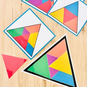Op de afbeelding: Meerdere vierkante en driehoekige kaarten met geometrische patronen bestaande uit kleinere driehoeken in levendige kleuren zoals rood, geel, groen, blauw, paars en oranje. Een losse, effen rode driehoek ligt aan de linkerkant.