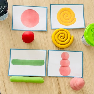 Könnte beinhalten: Vier rechteckige Aktivitätskarten mit Abbildungen eines roten Kreises, einer gelben Spirale, eines grünen Zylinders und eines dreistufigen rosa Stapels. Passende Knetmasseformen in Rot, Gelb, Grün und Rosa liegen neben den Karten.