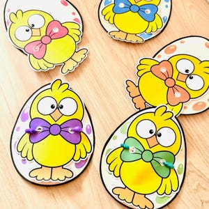Può includere: Quattro forme di uova in carta fustellata con pulcini gialli dei cartoni animati con occhi grandi e papillon colorati. Ogni pulcino è centrato su uno sfondo a forma di uovo con vari motivi. Alcuni papillon sono fissati con piccoli pezzi di filo.
