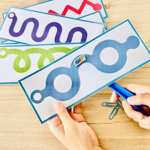 Puede incluir: Un conjunto de tarjetas rectangulares blancas con varios caminos de colores, incluyendo patrones de zigzag rojo, ondulado morado, bucles verdes y circulares azules. Un pequeño recorte de caracol está unido a la tarjeta azul, que es trazada por una herramienta de plástico azul.