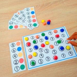 Könnte beinhalten: Ein Set aus pädagogischen Zählkarten und einem großen rechteckigen Arbeitsblatt mit nummerierten Kreisen. Das Material umfasst kleine, bunte, flauschige Pompons in Gelb, Orange, Rot, Blau und Grün sowie eine weiße Kunststoffpinzette.