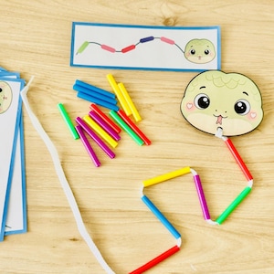 以下が含まれることがあります： 子供向けの知育玩具セット。紙製のヘビの頭の切り抜き、白い紐、様々な色のプラスチック製ストローのパーツが含まれています。長方形のカードには、紐とストローを使って再現するための色のパターンが描かれています。