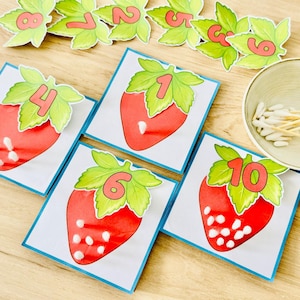 Könnte beinhalten: Ein Set aus Lernkarten mit Illustrationen roter Erdbeeren mit grünen Blättern und Zahlen von 1 bis 10. Jede Karte zeigt eine bestimmte Zahl, und weiße Wattestäbchen sind auf den Erdbeeren platziert, um die Samen darzustellen.