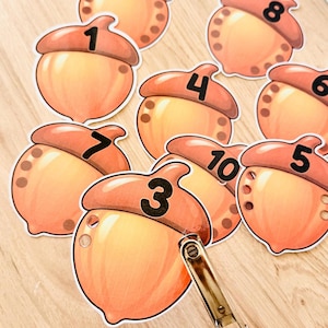 Könnte beinhalten: Eine Sammlung von ausgestanzten Eicheln aus Papier in Orange- und Brauntönen, die jeweils mit einer schwarzen Zahl von eins bis zehn bedruckt sind. Ein Metall-Locher liegt auf der Eichel mit der Nummer drei und zeigt kleine gestanzte Löcher.