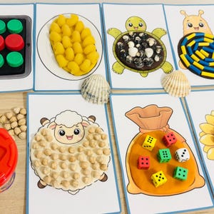 Puede incluir: Un conjunto de tarjetas de actividades educativas con ilustraciones de una bandeja, un plato de pasta amarilla, una tortuga, un caracol, una oveja y un saco. Las tarjetas están decoradas con garbanzos, dados, conchas y tapones de plástico.