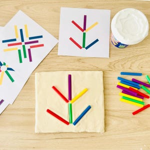 Puede incluir: Un cuadrado de masa para modelar color hueso con un patrón geométrico hecho de pequeños palitos de plástico de colores. Cerca hay tarjetas con patrones impresos, un recipiente de plástico blanco y una colección de palitos sueltos rojos, azules, verdes, amarillos y morados.