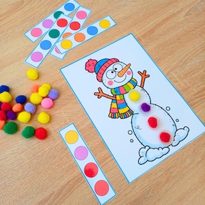 May include: A children's activity sheet featuring a cartoon snowman illustration with a blue hat and colorful striped scarf. The set includes several paper strips with colored circles and a collection of small, multi-colored pom-poms.