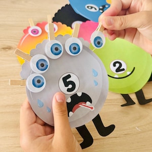 Puede incluir: Un conjunto de recortes de papel circulares con forma de monstruo en varios colores, incluyendo gris, verde y naranja. Cada monstruo presenta un número grande, una boca con dientes y pinzas de madera para ropa sujetas a los bordes.