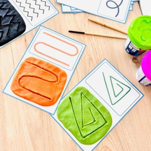 May include: Educational activity cards featuring printed line patterns paired with orange and green modelling clay. The clay is pressed onto the cards to match the printed shapes, accompanied by two wooden styluses and two plastic containers.