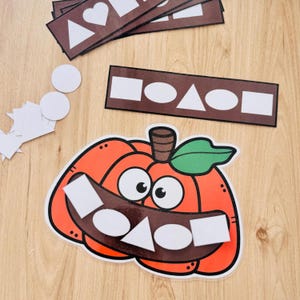 Pode incluir: Um recorte de abóbora laranja laminado com uma folha verde e um caule castanho, apresentando uma boca castanho-escura com formas geométricas brancas, incluindo um quadrado, um oval e um triângulo. Inclui tiras castanhas com formas brancas.