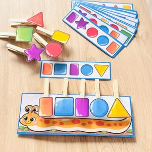 May include: Educational matching game featuring wooden clothespins with colorful geometric shapes including triangles, squares, circles, and stars. The set includes printed cards with matching shapes and a cartoon caterpillar illustration for shape recognition.