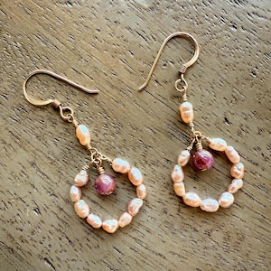 May include: A pair of gold-toned hoop earrings. Each earring features a circle of small, peach-colored pearls and a single, round, pink gemstone. The earrings have a simple hook closure.