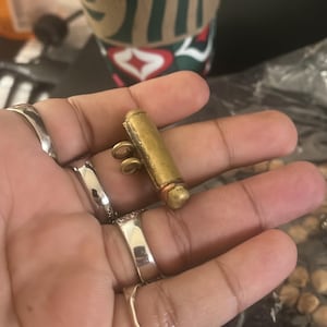 May include: A small, cylindrical brass pendant with a loop for hanging. The pendant has a slightly aged appearance, with a copper-colored band near one end. Several silver rings adorn the fingers holding the pendant. A coffee cup is visible in the background.