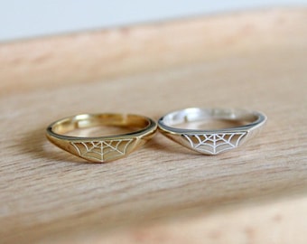 Könnte beinhalten: Zwei Ringe, einer goldfarben und einer silberfarben, jeweils mit einem filigranen Spinnennetz-Design. Die Ringe liegen auf einer Holzoberfläche, die ihre komplizierten Details und kontrastierenden Metalloberflächen hervorhebt. Der goldfarbene Ring hat ein warmes, poliertes Aussehen, während der silberfarbene einen kühlen, hellen Kontrast bietet.