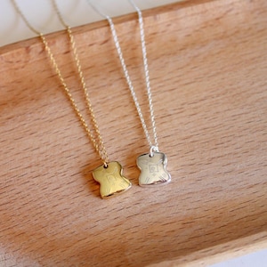 May include: Two necklaces with thin chains, one in gold tone and one in silver tone. Each necklace features a small, smooth, irregular square-shaped pendant with rounded corners and engraved geometric symbols on the front surface.