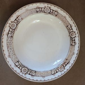 May include: A white ceramic plate with a brown floral and geometric border design. The plate has a wide rim with alternating floral and geometric patterns. The center of the plate is plain white.