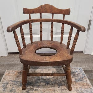 May include: Antique wooden child's potty chair with a round seat opening. The chair has a curved back and armrests, and is constructed with turned spindles and legs. The wood has a warm, aged patina.