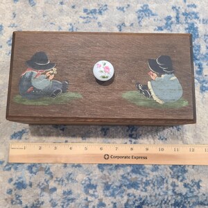 May include: A wooden box with a floral knob and painted illustrations of children in hats. The box is brown and rectangular, with a ruler placed below it. The children are depicted in blue and gray clothing.