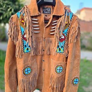 May include: A brown suede jacket with fringe and beaded details. The jacket features colorful beadwork on the shoulders and pockets, with a Native American-inspired design. The jacket has a collar and button closure, with the text "Real Leather" on the label.