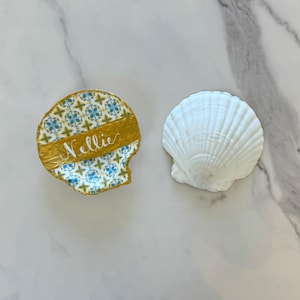 May include: Two decorative seashells. One is painted with a gold band and a blue and yellow pattern, with the name "Nellie" in cursive. The other is a natural white scallop shell.