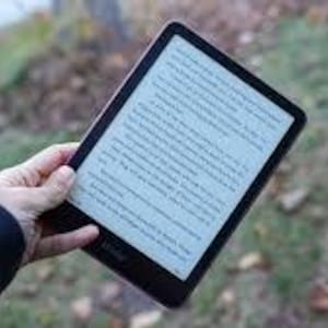 May include: A black e-reader with a white screen displaying text is held in a hand. The device has a thin bezel and a dark border. The background is blurred, showing foliage.
