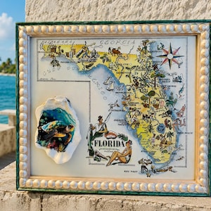 May include: A framed decorative map of Florida featuring illustrated landmarks and figures, accented by a three-dimensional oyster shell attached to the left side, which displays a colourful painted sea turtle swimming in blue water.